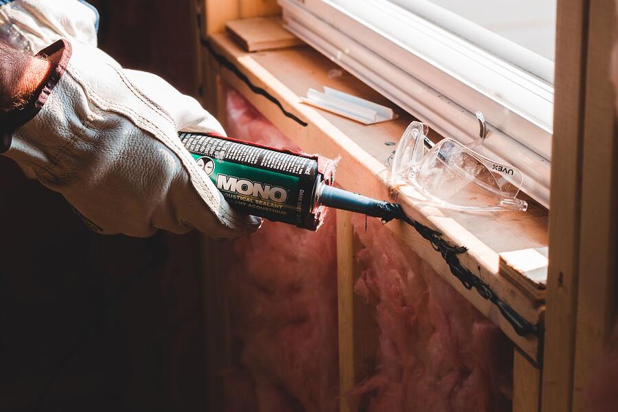 A gloved hand running a bead of sealant along a gap at a window frame