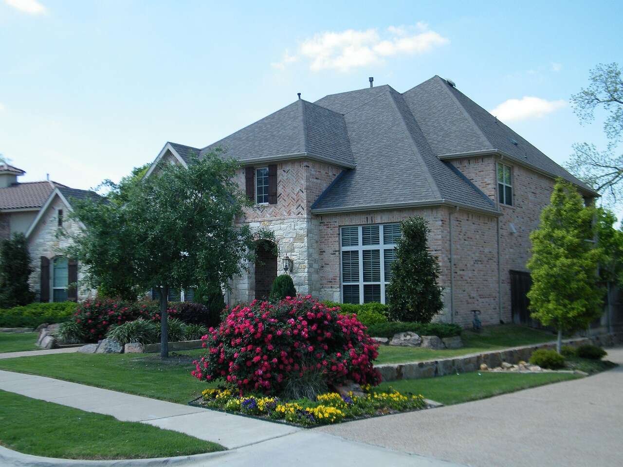 A two-story brick house with a porch and flower beds on a wide lot