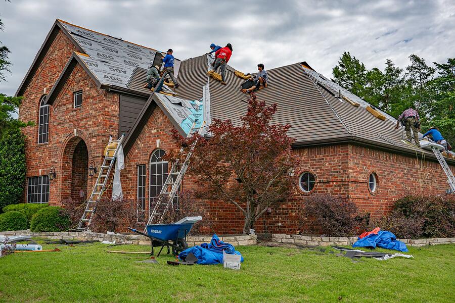 Roofers on ladders at the eave of a brick Tudor-style house, with shingle bundles on the lawn